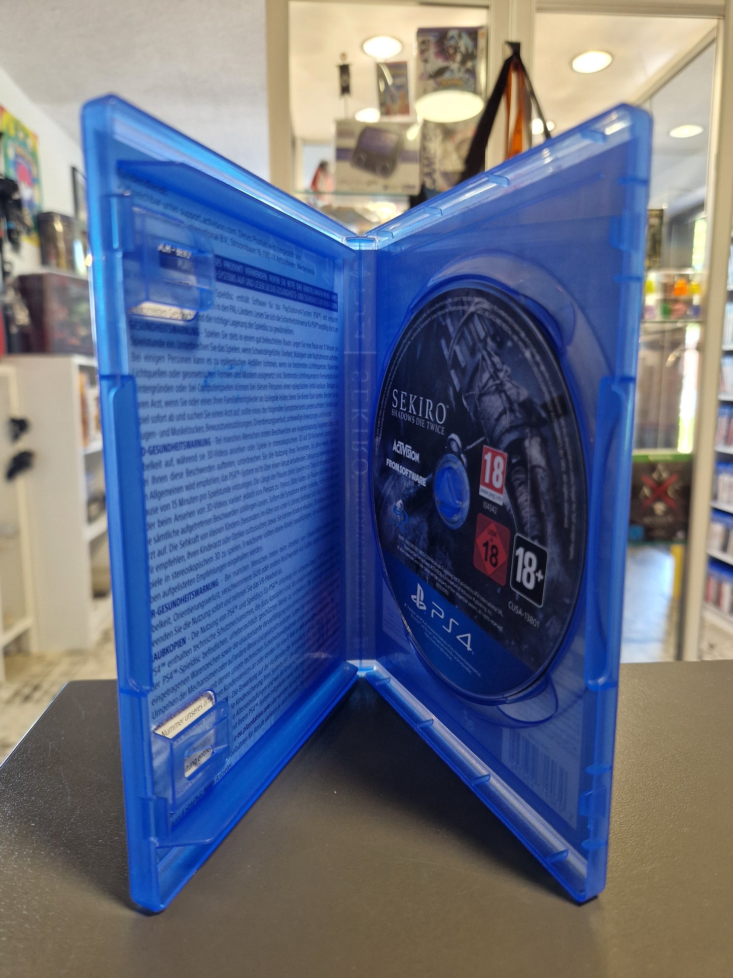 Die geöffnete, blaue PS4 Verpackung zeigt die unbeschädigte, kratzerfreie Sekiro: Shadows Die Twice Spieldisc, eingelegt auf der rechten Seite. Das Innencover ist mit Hinweistexten versehen.