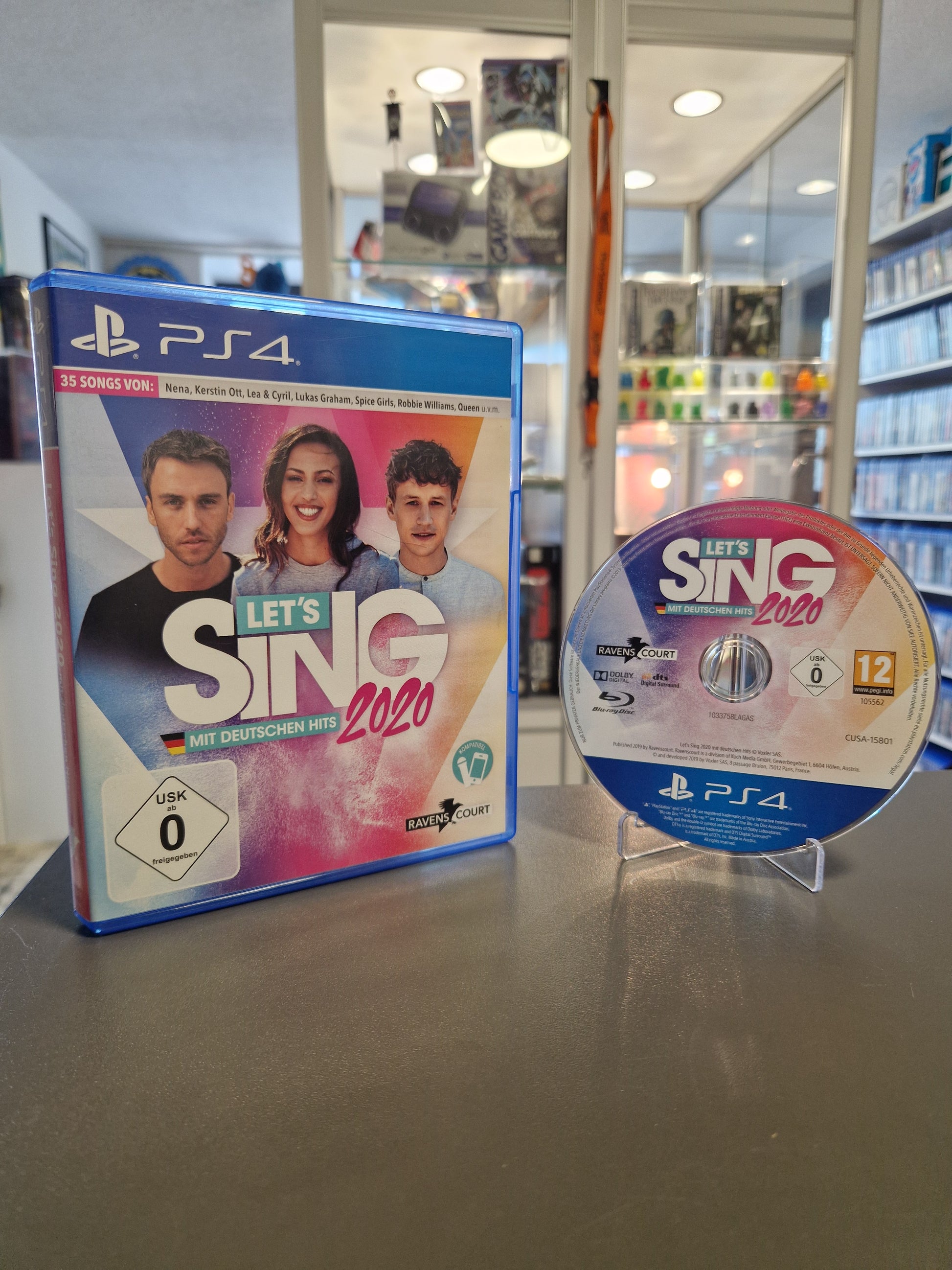 Die PlayStation 4 Spielehülle von Let's Sing 2020 mit deutschen Hits neben der passenden Blu-ray Disc auf einem Acrylständer. Auf dem Cover ist ein USK 0 Logo zu sehen.