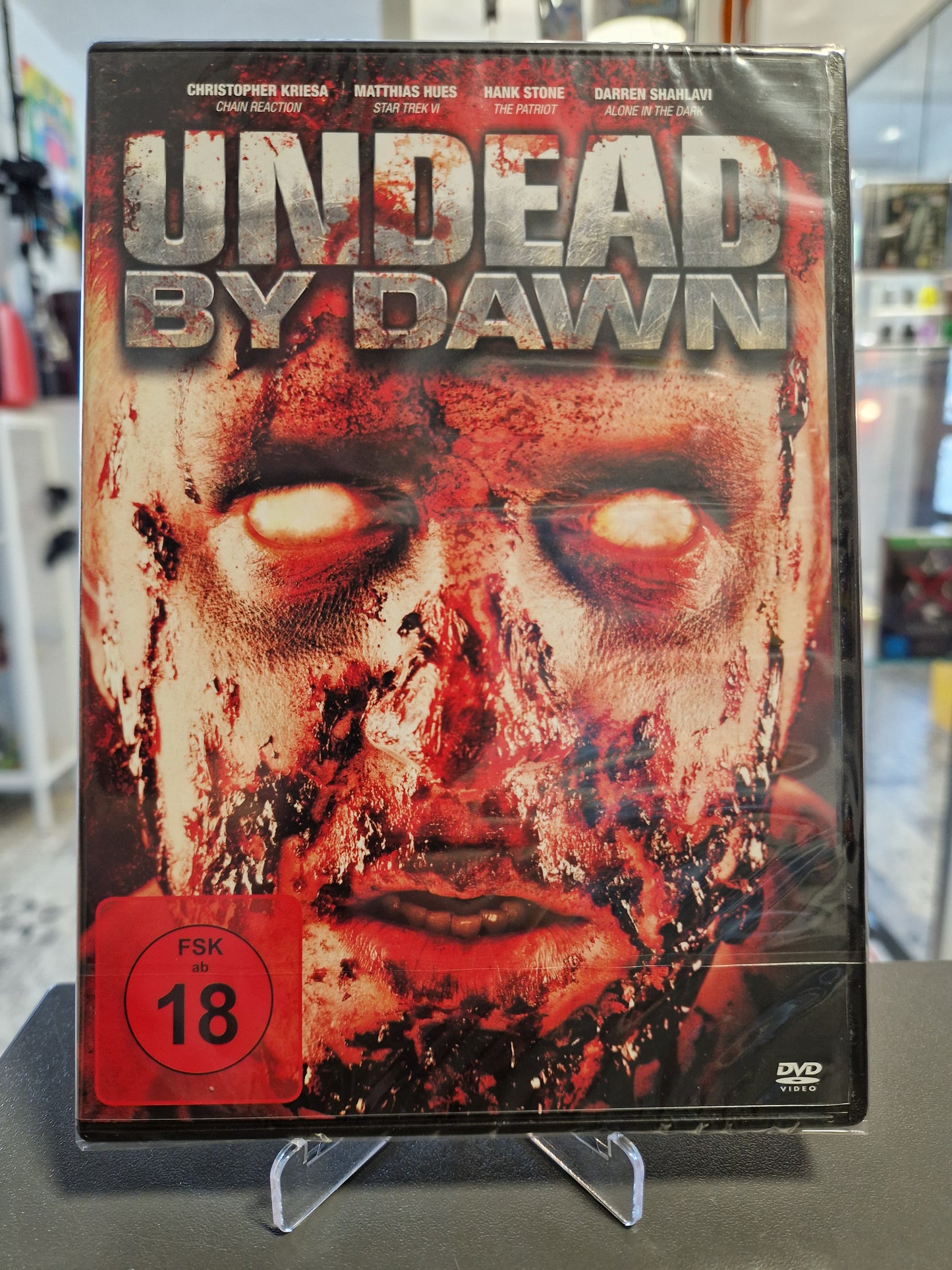 Originalverpackte und in Folie eingeschweißte DVD des Horrorfilms Undead by Dawn mit rotem FSK 18 Logo. Das Cover zeigt ein blutüberströmtes Zombiegesicht mit weiß leuchtenden Augen.