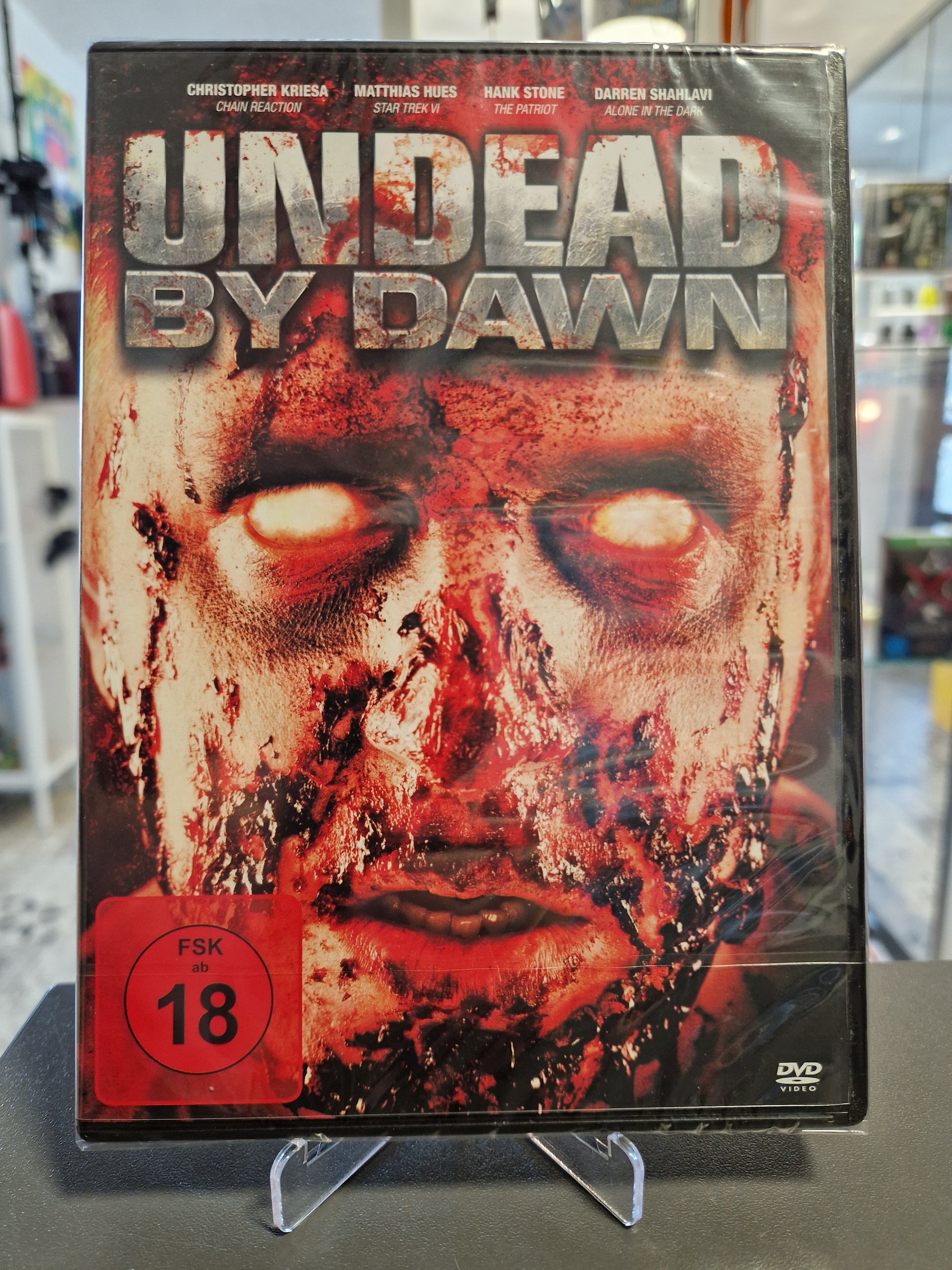 Originalverpackte und in Folie eingeschweißte DVD des Horrorfilms Undead by Dawn mit rotem FSK 18 Logo. Das Cover zeigt ein blutüberströmtes Zombiegesicht mit weiß leuchtenden Augen.