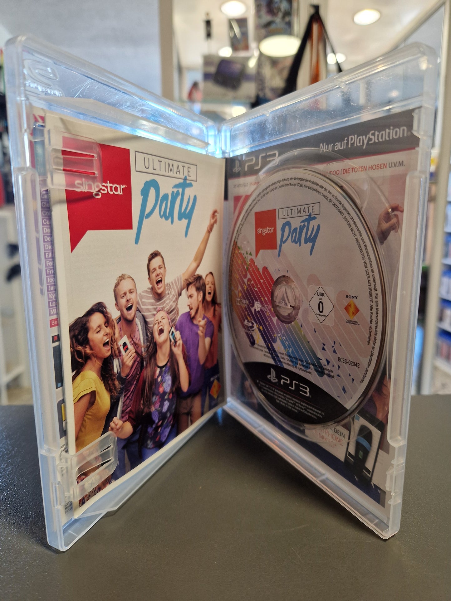 Die geöffnete, transparente PS3 Hülle zeigt das originale SingStar: Ultimate Party Begleitheft auf der linken Seite und die saubere, kratzerfreie Spieldisc mit Musikdesign auf der rechten Seite.