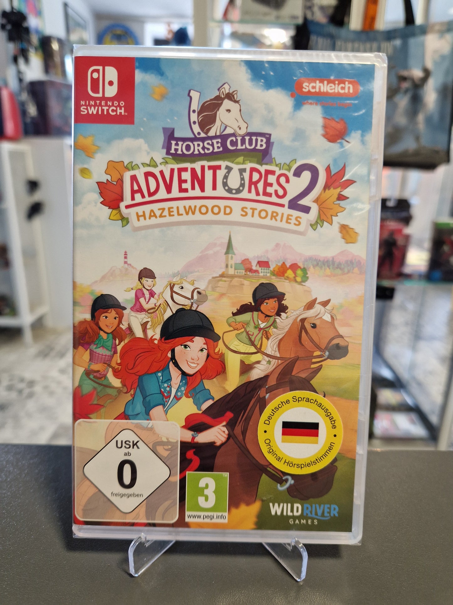 Vorderseite der originalverpackten Hülle von Horse Club Adventures 2 Hazelwood Stories für Nintendo Switch mit den reitenden Schleich-Charakteren, USK 0 Logo und gelbem Sticker zur deutschen Sprachausgabe.