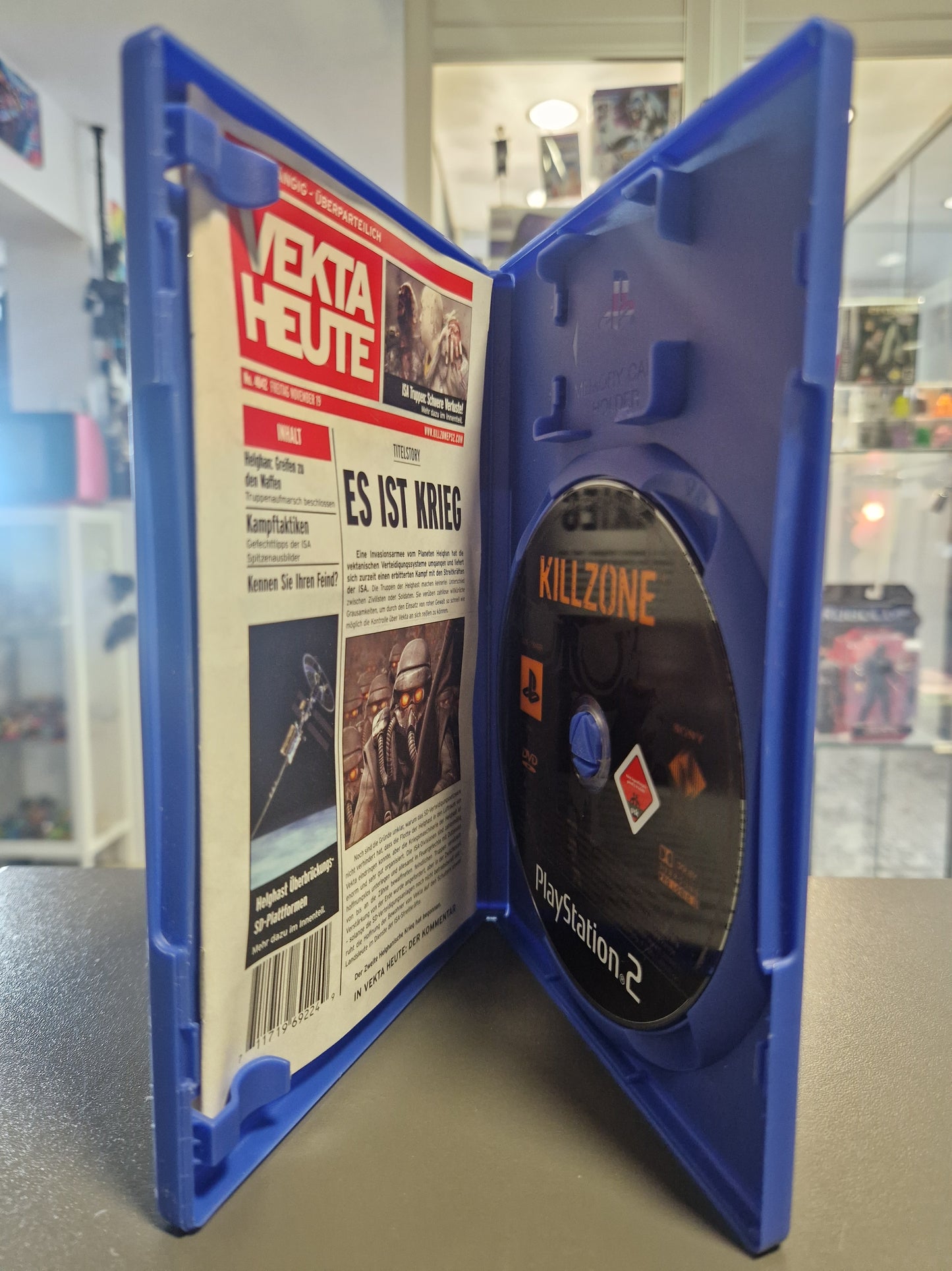 zeigt die geöffnete blaue PS2-Hülle mit der originalen, schwarzen Spiele-Disc auf der rechten Seite und der als Zeitung „Vekta Heute“ designten Spieleanleitung auf der linken Seite.