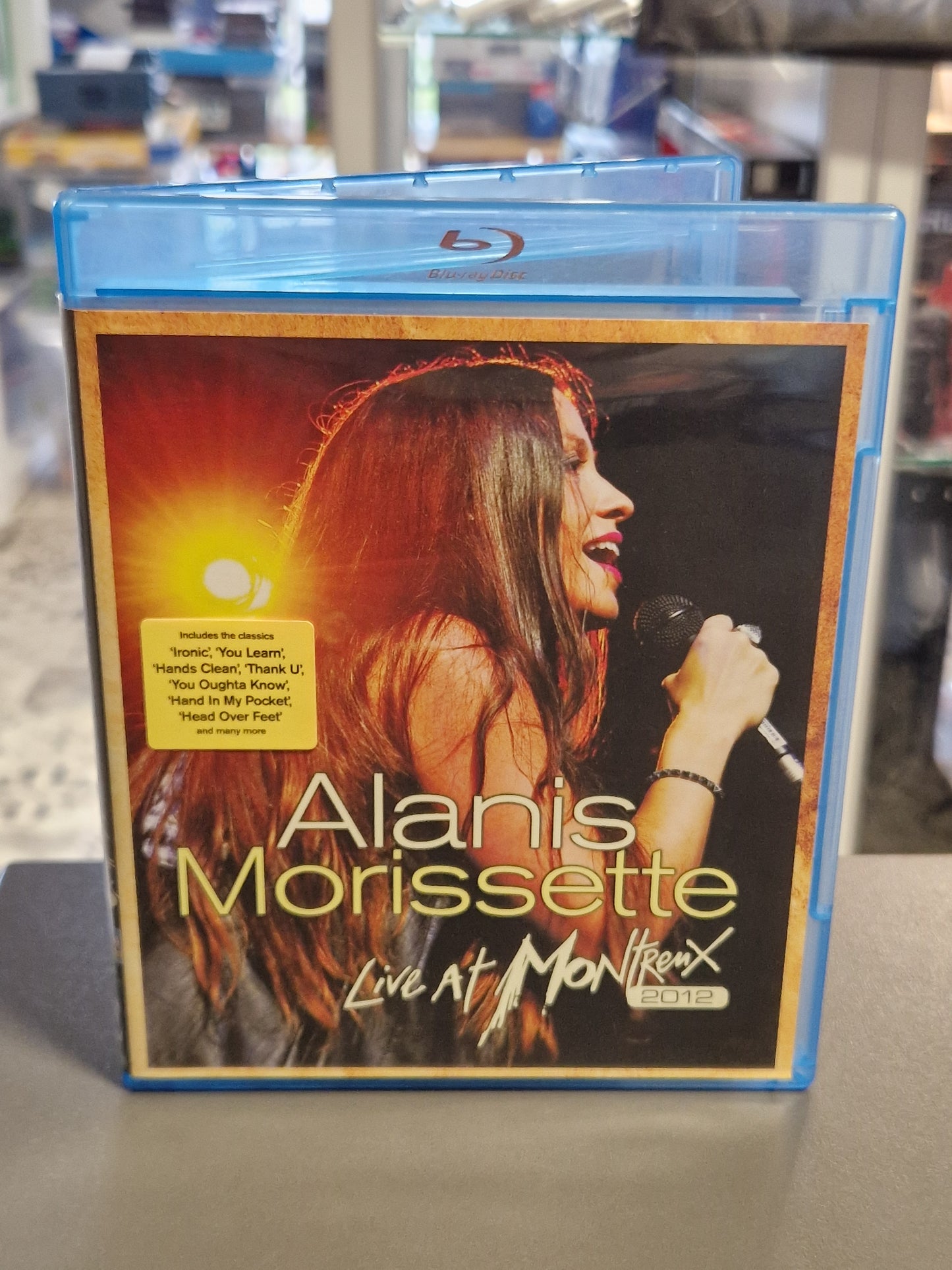 Vorderseite der Blu-ray Hülle von Alanis Morissette Live at Montreux 2012 mit einem Konzertfoto der Sängerin am Mikrofon und einem gelben Info-Sticker zu den enthaltenen Klassikern.