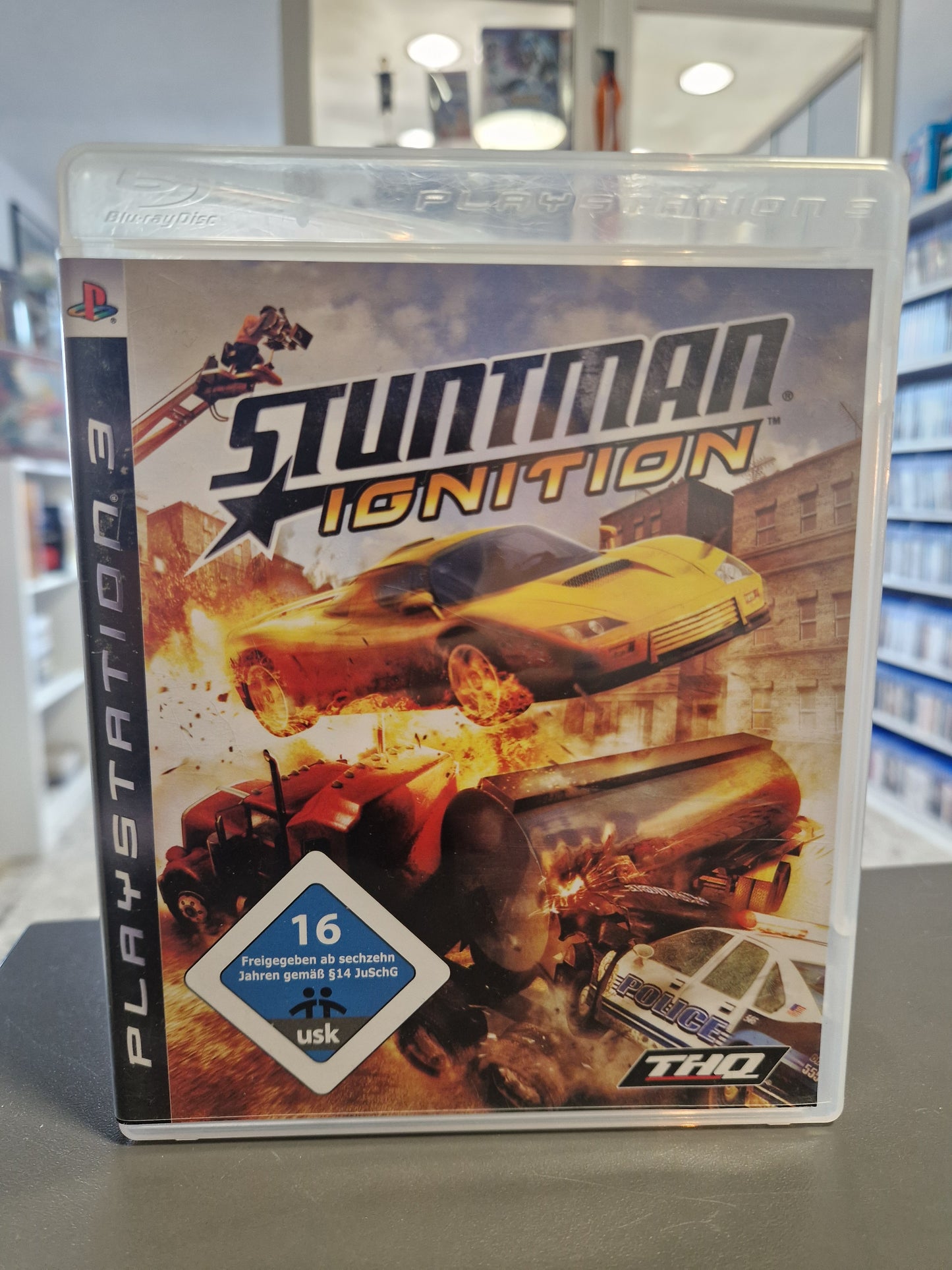 PS3-Cover von „Stuntman: Ignition“ zeigt ein brennendes Auto, das spektakulär durch die Luft fliegt, während im Hintergrund Filmkameras und Explosionen zu sehen sind.