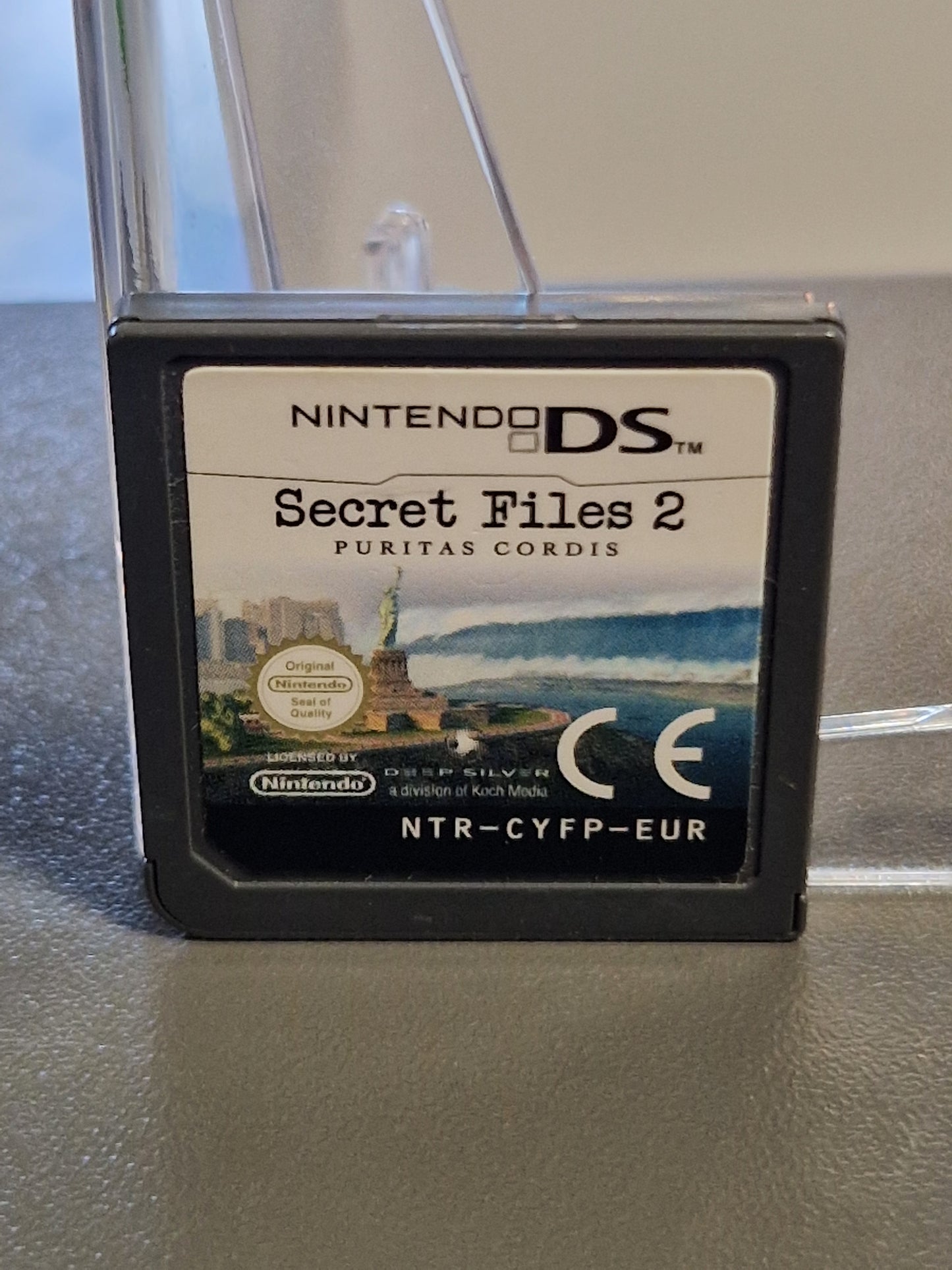 Originales Nintendo DS Spielmodul von Secret Files 2 Puritas Cordis auf einem Ständer. Das Label ist sauber und zeigt das CE-Zeichen sowie die Freiheitsstatue im Hintergrund.