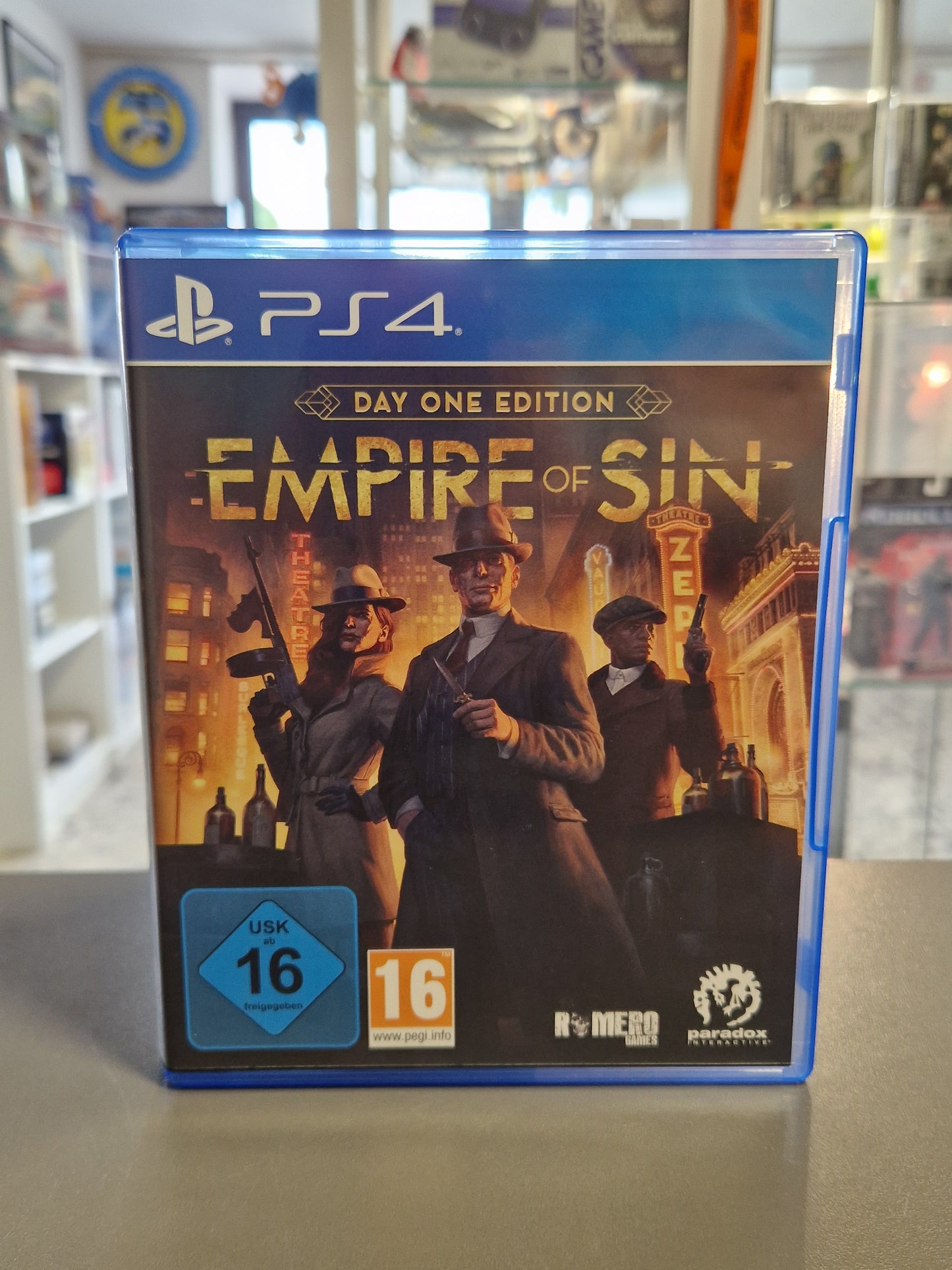 Vorderseite der originalen PS4-Hülle von „Empire of Sin - Day One Edition“ mit dem Artwork von drei eleganten Mafiosi im Stil der 1920er-Jahre vor einer nächtlichen Stadtkulisse sowie den Logos von Romero Games und Paradox.