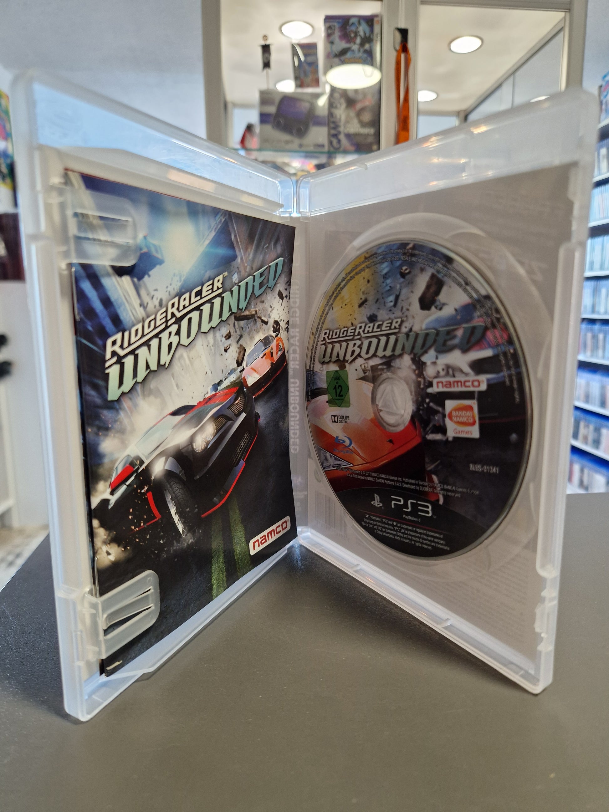 Geöffnete transparente PlayStation 3 Hülle, die die eingelegte Ridge Racer Unbounded Spiele-Disc auf der rechten Seite und das farbige Handbuch auf der linken Seite zeigt.