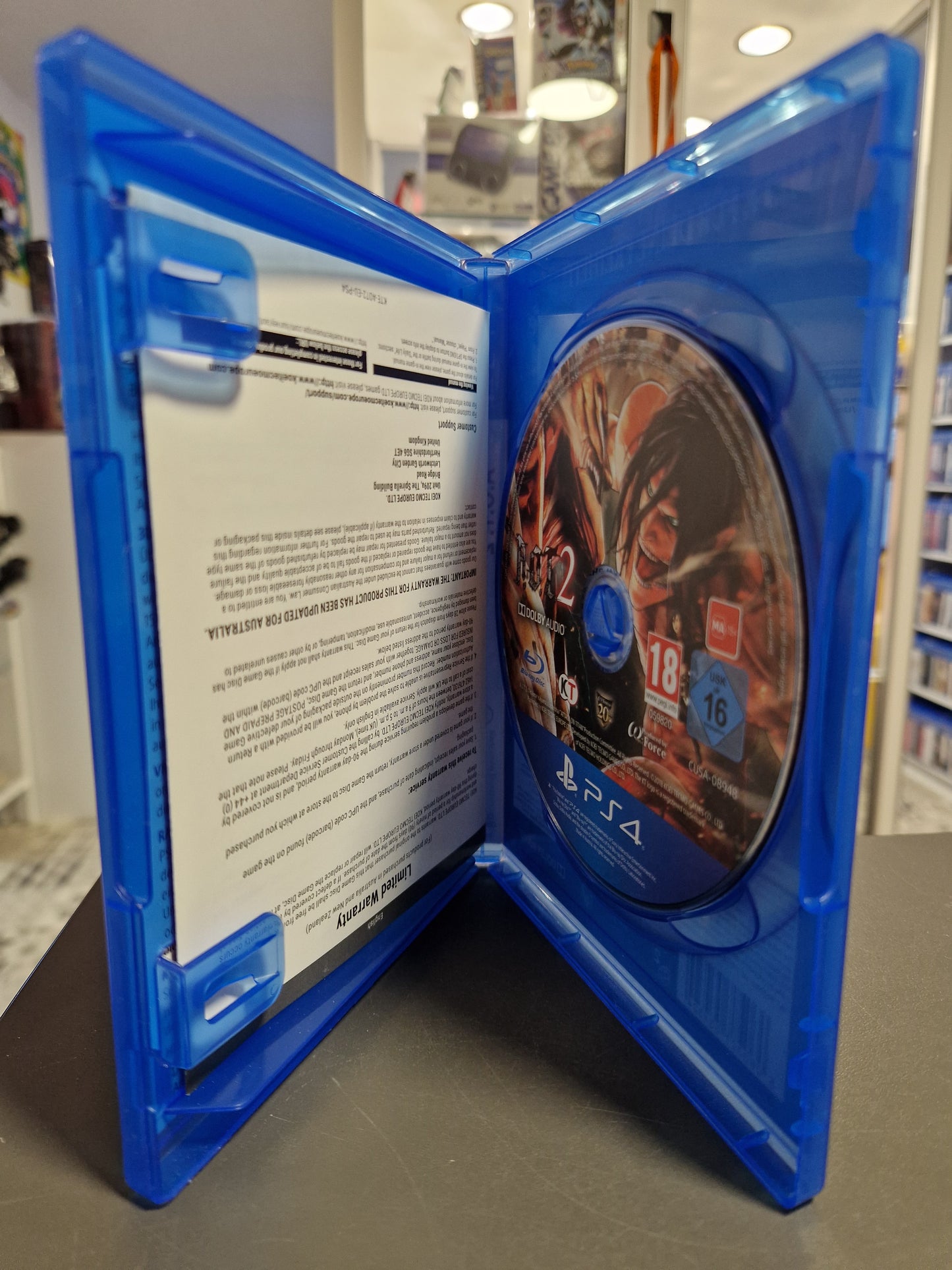 Innenansicht der A.O.T. 2 PS4-Hülle. Die Spiel-Disc zeigt ein Motiv von Eren in Titanen-Form. Ein beiliegendes Informationsblatt ist auf der linken Seite zu sehen.