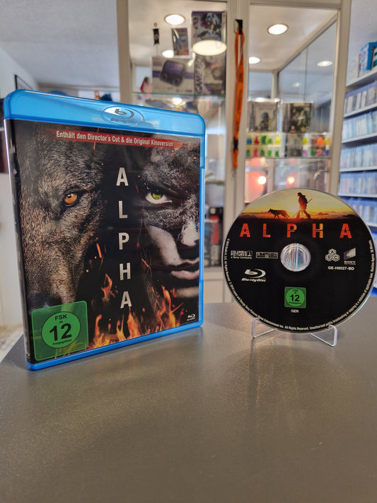 Die blaue Blu-ray-Hülle des Films Alpha steht neben der zugehörigen Disc auf einem transparenten Ständer. Das Cover zeigt das geteilte Gesicht eines Wolfes und eines Jägers im Eiszeit-Look mit grünem FSK 12 Logo.