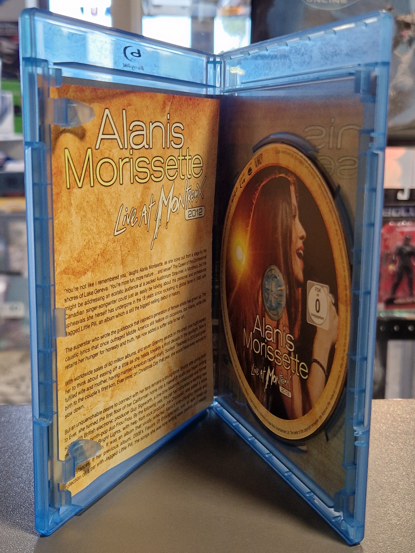 Geöffnete Blu-ray Hülle von Alanis Morissette Live at Montreux 2012 mit der goldfarbenen Konzert-Disc auf der rechten Seite und dem aufgeschlagenen Inlay-Booklet mit englischem Begleittext auf der linken Seite.