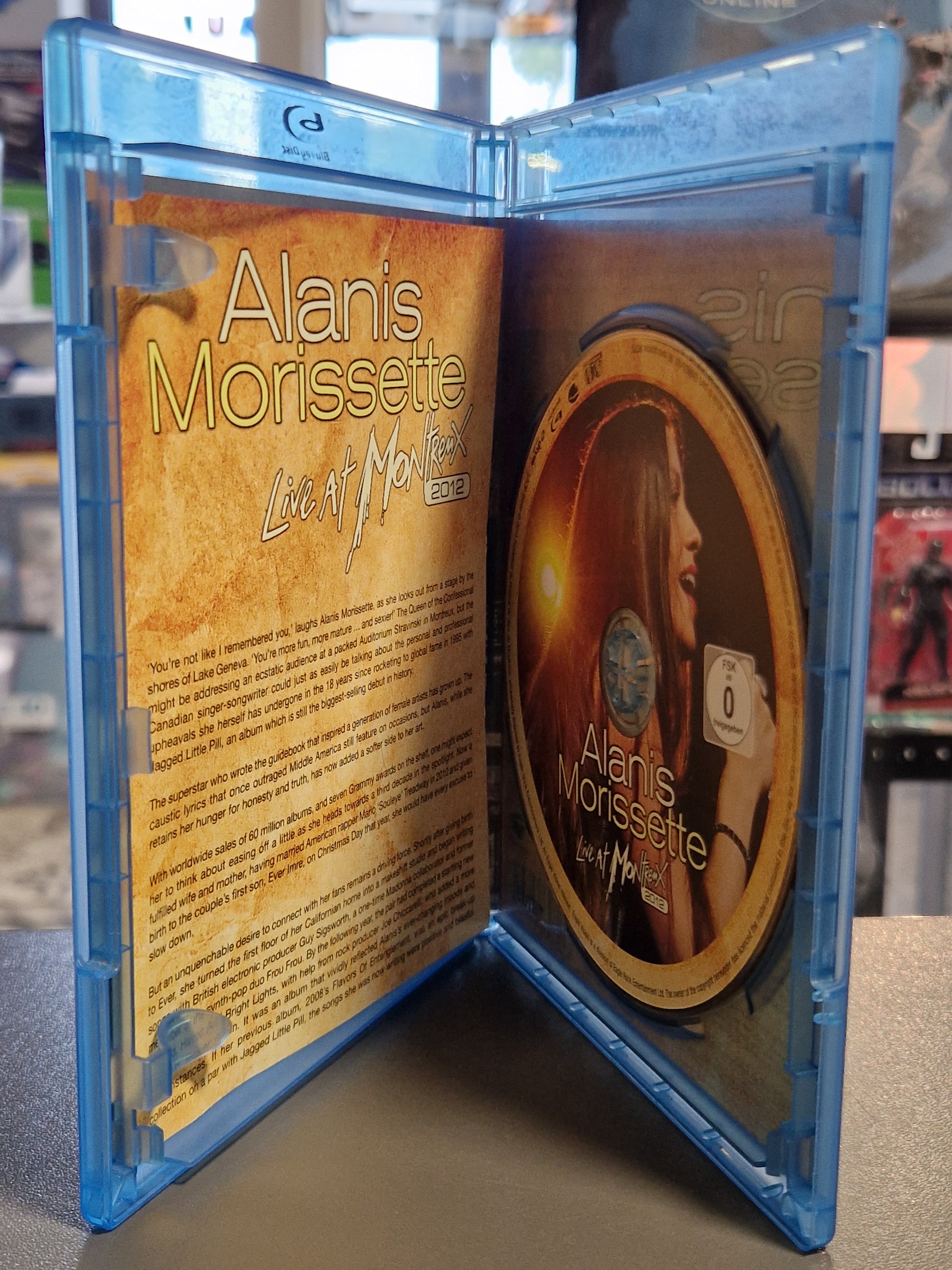 Geöffnete Blu-ray Hülle von Alanis Morissette Live at Montreux 2012 mit der goldfarbenen Konzert-Disc auf der rechten Seite und dem aufgeschlagenen Inlay-Booklet mit englischem Begleittext auf der linken Seite.