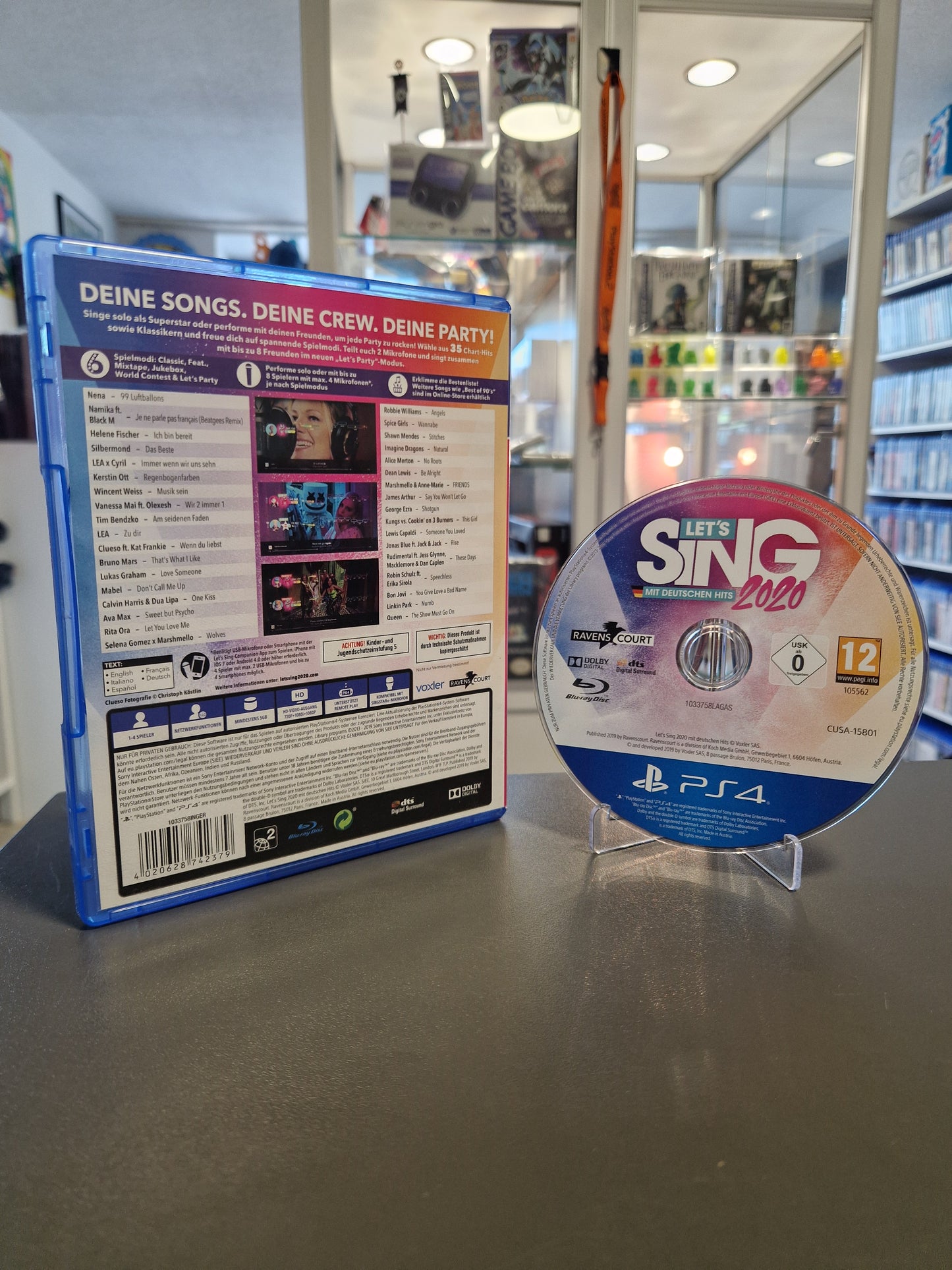 Die Rückseite der PS4 Hülle von Let's Sing 2020 mit der vollständigen Songliste, Feature-Beschreibungen und dem Barcode. Daneben steht die saubere Spiele-Disc.