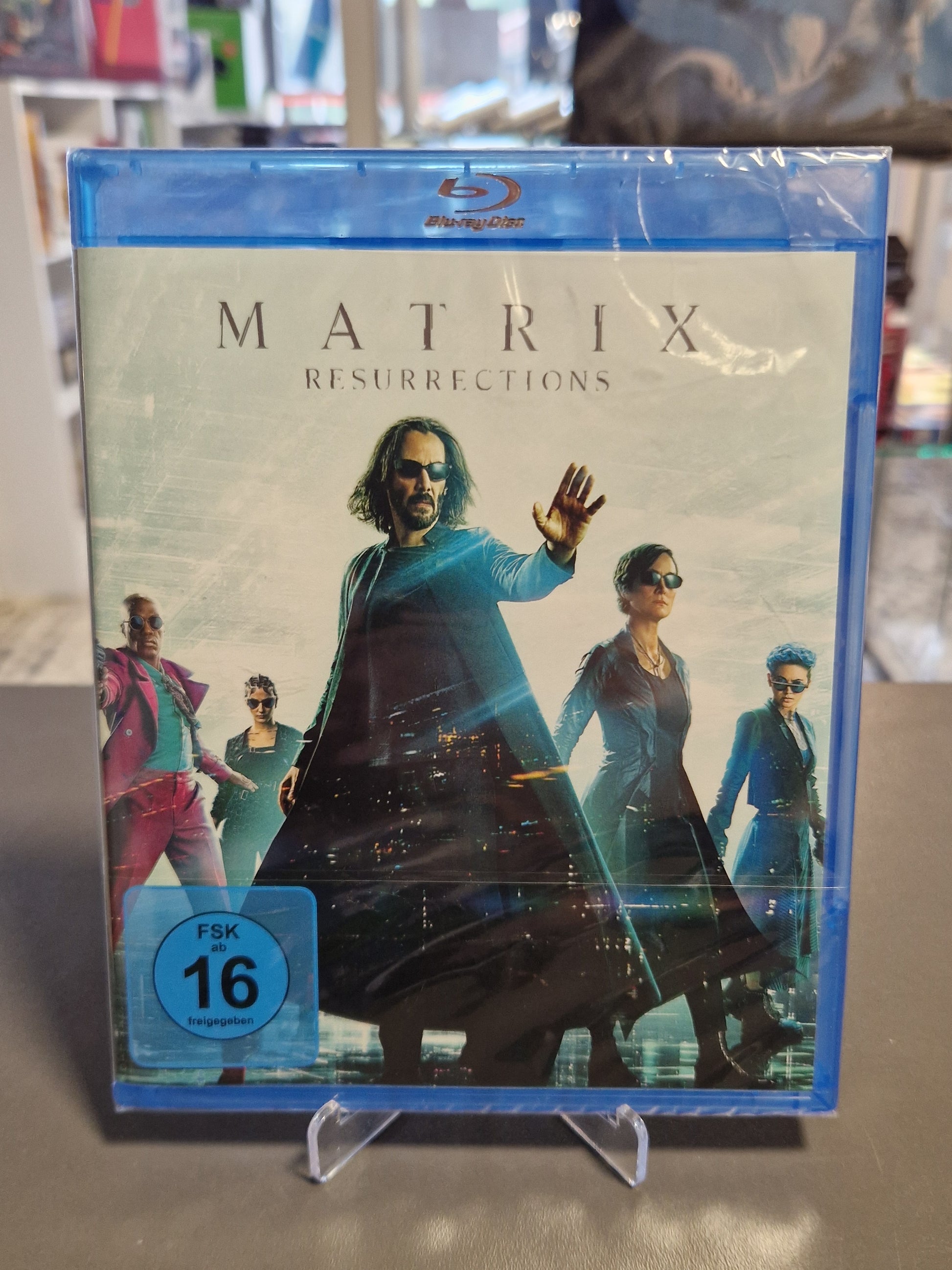 Vorderseite der originalverpackten Hülle von Matrix Resurrections auf Blu-ray mit Keanu Reeves als Neo, Carrie-Anne Moss als Trinity, blauem FSK 16 Logo und sichtbarem kleinen Riss oben rechts an der Hülle.