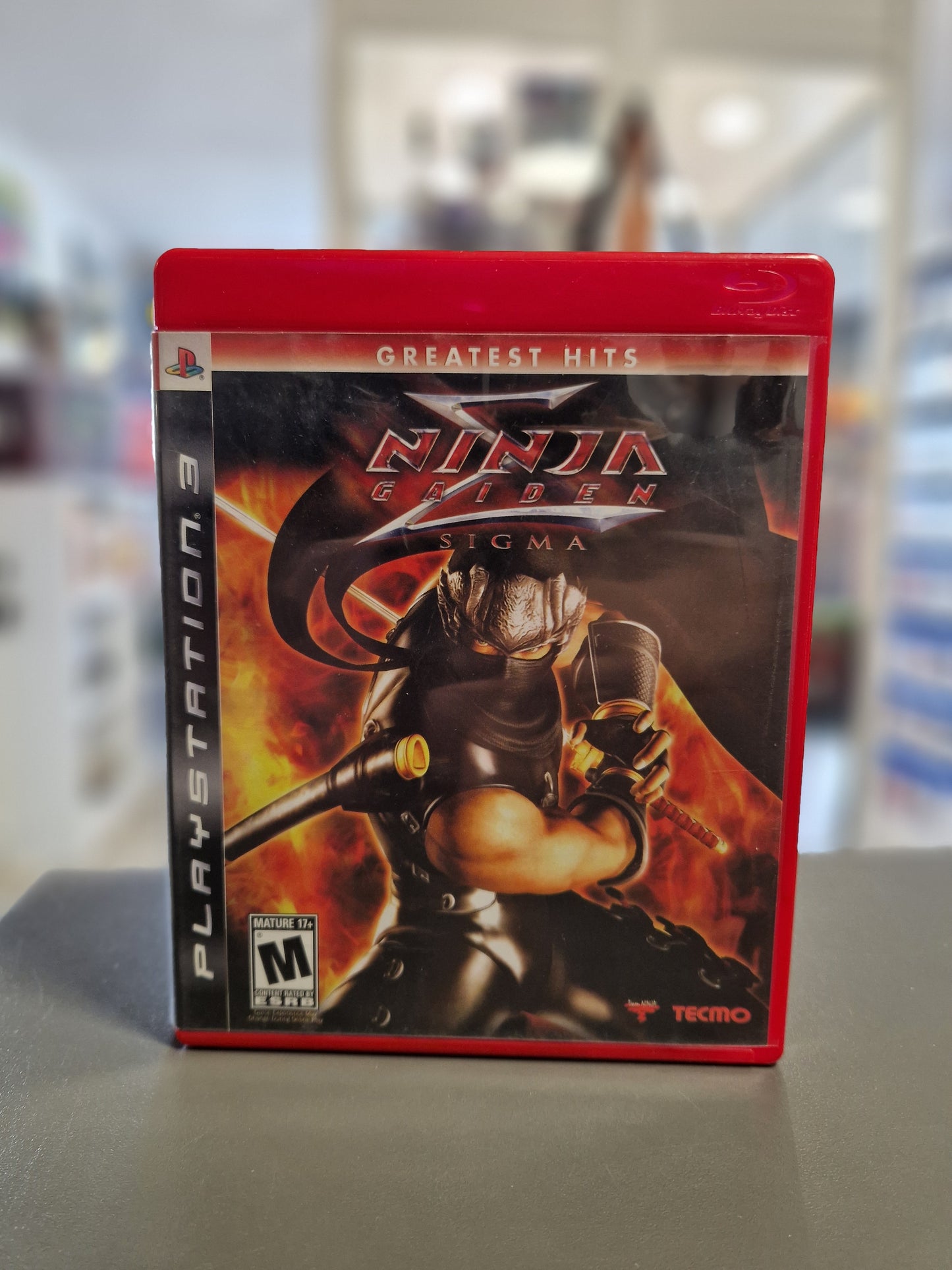 Die rote PlayStation 3 Greatest Hits Spielehülle von Ninja Gaiden Sigma (US-Import). Das Cover zeigt den Ninja Ryu Hayabusa in Kampfpose vor einer feurigen Kulisse sowie das ESRB M-Logo.